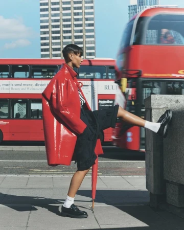 Fashionable woman in red coat poses in city with double-decker bus. Dark hair, black shorts, and umbrella visible in an urban setting.