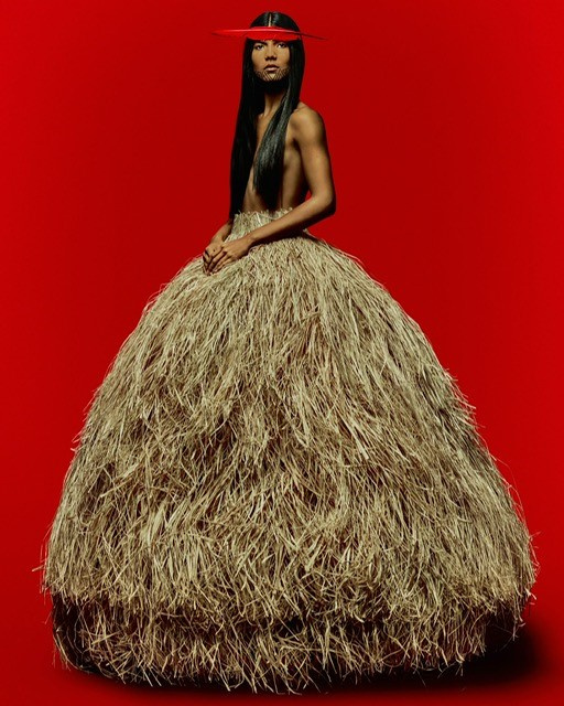 Woman in textural beige dress with red bar across eyes poses against a red background in a studio shot. Editorial fashion.