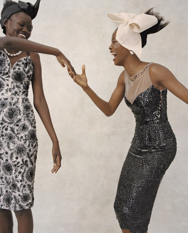 Two dark-skinned women in formal black and white dresses and headpieces laugh and touch hands in a light neutral-toned setting.