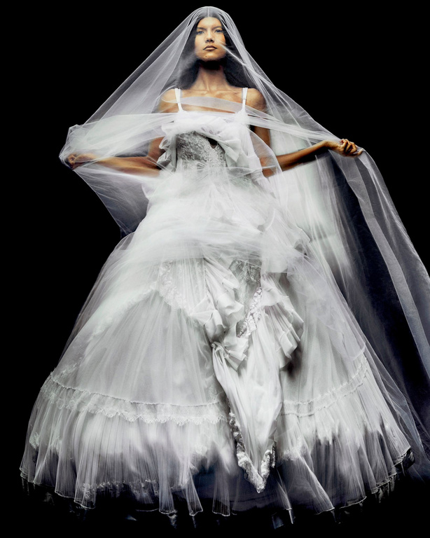 A woman poses in a white couture gown with a long sheer veil against a stark black background in a fashion photography studio.