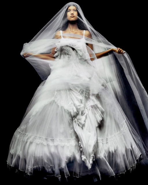 A woman poses in a white couture gown with a long sheer veil against a stark black background in a fashion photography studio.