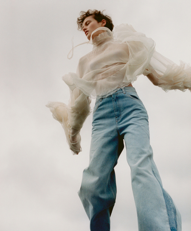 Model in sheer ruffled white blouse and light blue jeans shot from a low worm's-eye angle against an overcast sky.