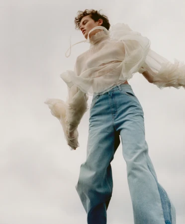 Model in sheer ruffled white blouse and light blue jeans shot from a low worm's-eye angle against an overcast sky.