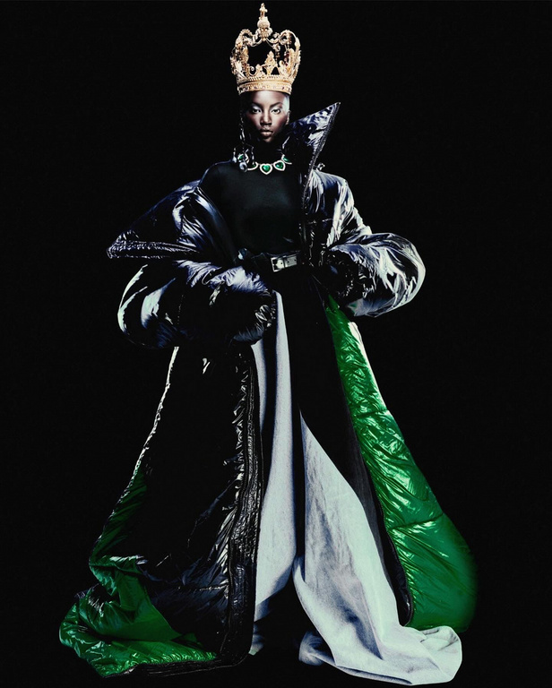 Portrait of African-American woman wearing crown, necklace, and large black, green, and white puffer jacket against a black backdrop.