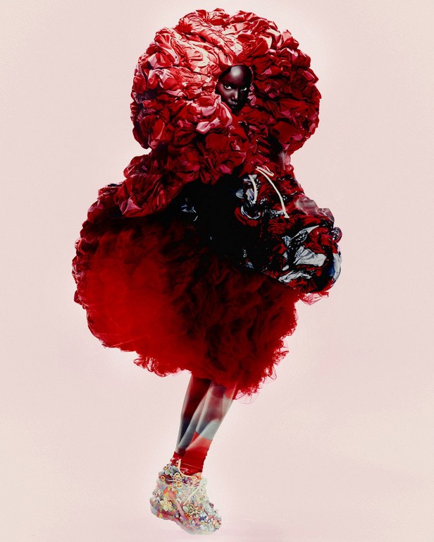 Runway fashion show featuring a Black female model wearing an elaborate, oversized red gown with embellished shoes in a studio setting.