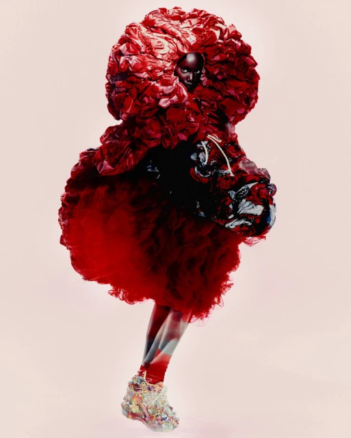Runway fashion show featuring a Black female model wearing an elaborate, oversized red gown with embellished shoes in a studio setting.
