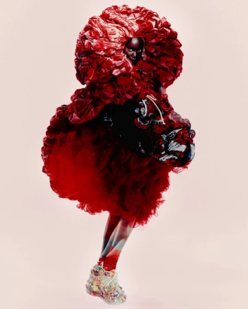 Runway fashion show featuring a Black female model wearing an elaborate, oversized red gown with embellished shoes in a studio setting.