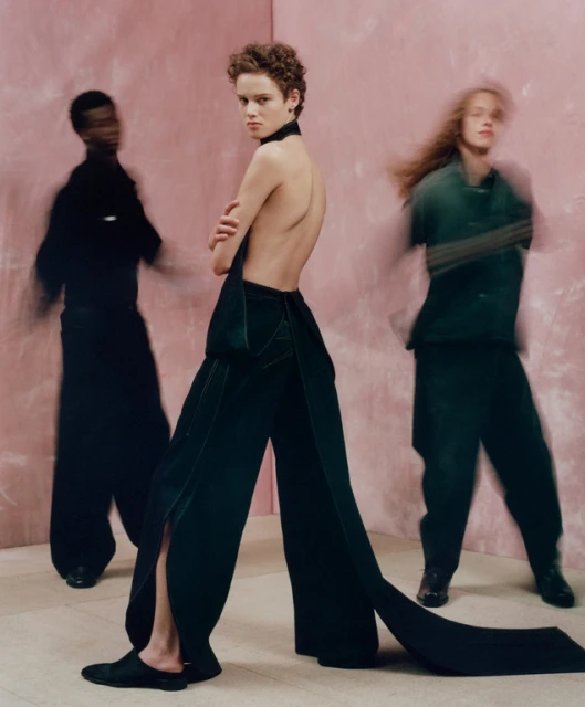 Three fashion models wearing dark clothing posing in front of a pink wall. One in focus looking over shoulder.