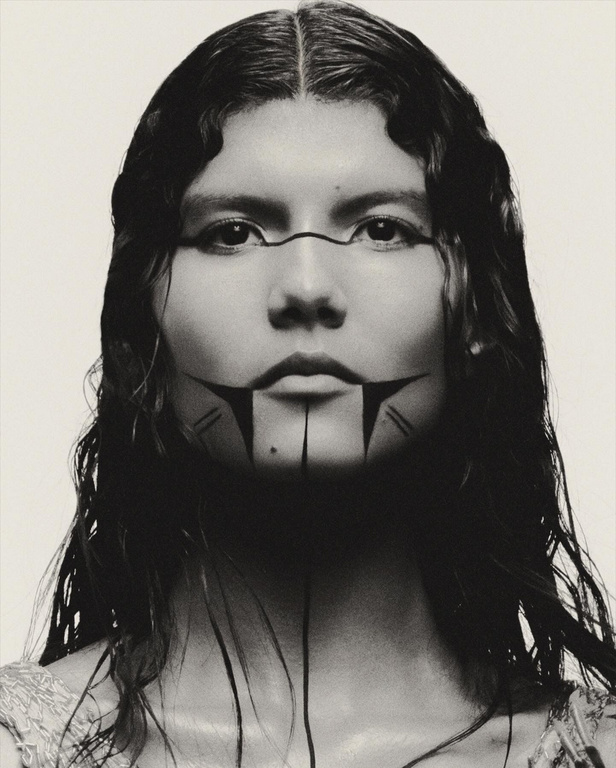 Close-up portrait of a woman with dark hair and painted facial markings, set against a plain white background in a studio setting.