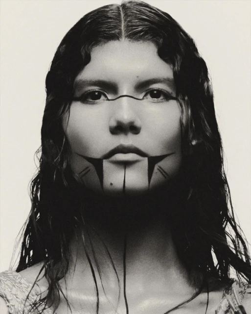 Close-up portrait of a woman with dark hair and painted facial markings, set against a plain white background in a studio setting.
