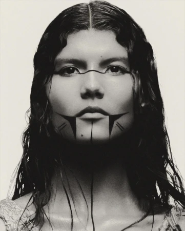 Close-up portrait of a woman with dark hair and painted facial markings, set against a plain white background in a studio setting.
