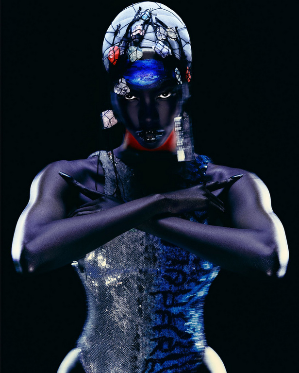 Striking portrait of a female figure with dark skin, distinctive headwear, dramatic makeup, crossed arms, and a shimmering bodysuit on a black backdrop.