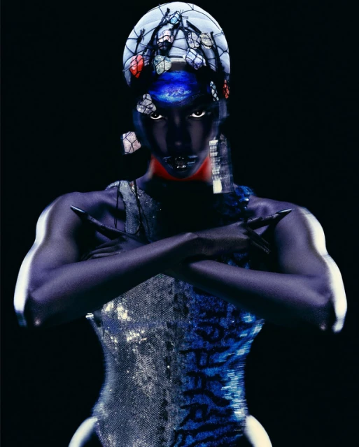 Striking portrait of a female figure with dark skin, distinctive headwear, dramatic makeup, crossed arms, and a shimmering bodysuit on a black backdrop.