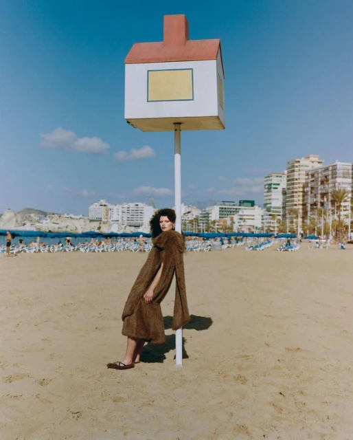 Brunette woman in a brown coat leans against a white post on a beach with buildings, people, and blue sky in the background.