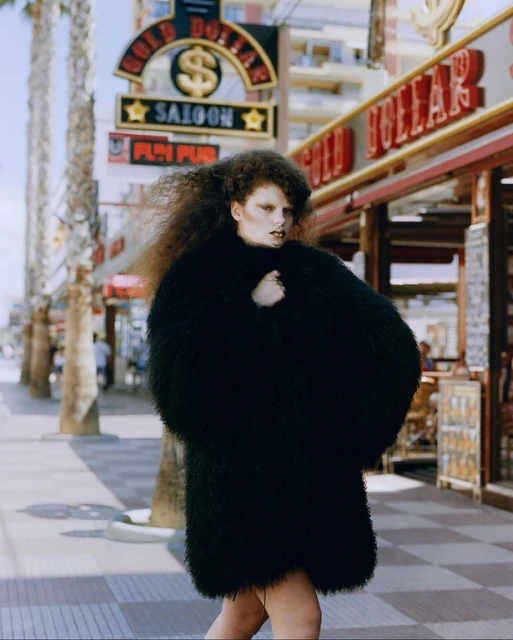 A woman walks on a sunny street wearing a large black coat with dark hair, posing near the Gold Dollar Saloon and storefronts.