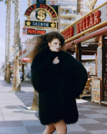 A woman walks on a sunny street wearing a large black coat with dark hair, posing near the Gold Dollar Saloon and storefronts.