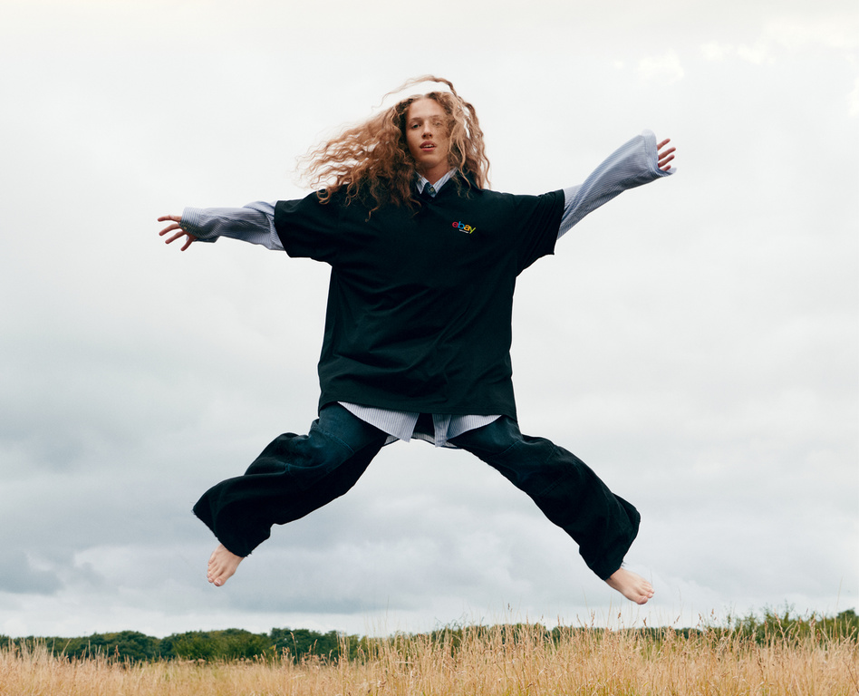 Woman with curly hair jumps barefoot outdoors, wearing a black shirt, jeans, and a gray collared shirt. She leaps against cloudy sky.