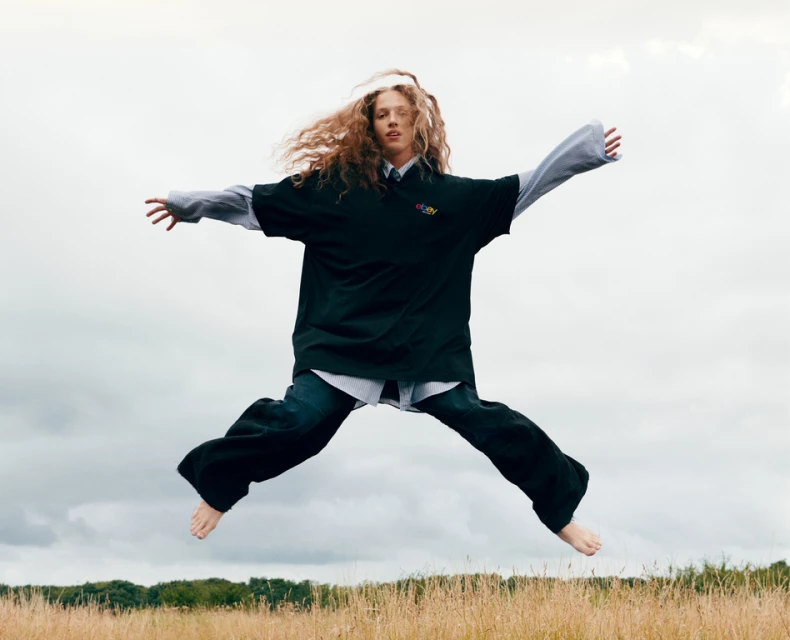 Woman with curly hair jumps barefoot outdoors, wearing a black shirt, jeans, and a gray collared shirt. She leaps against cloudy sky.