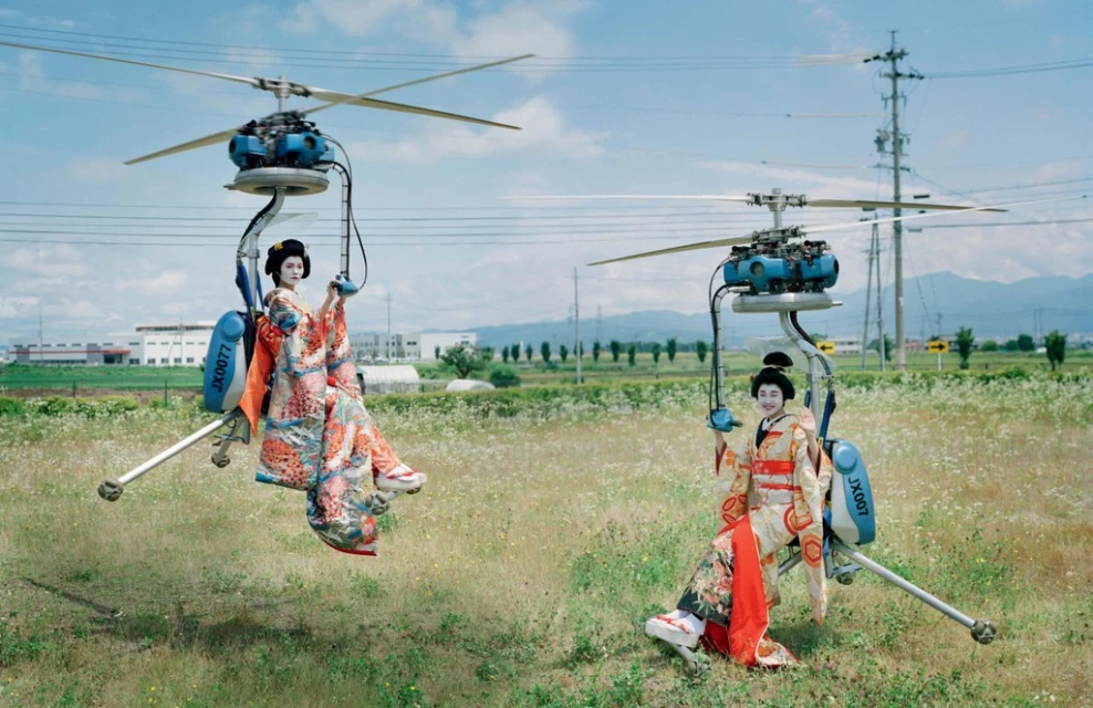 Two women in kimonos on personal helicopter seats hover over a grassy field under a blue sky with buildings and mountains visible.