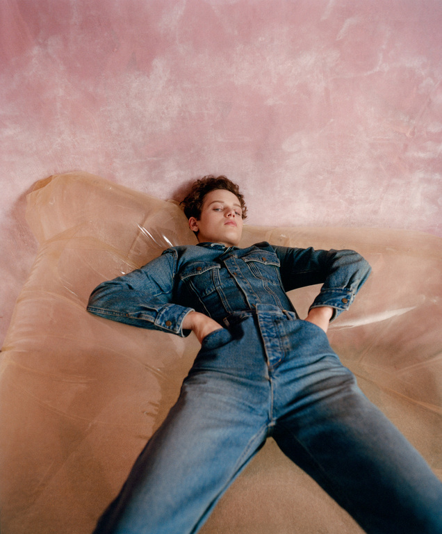 Young adult in a denim jumpsuit reclines on a clear inflated surface against a pink mottled background in a fashion-style studio portrait.
