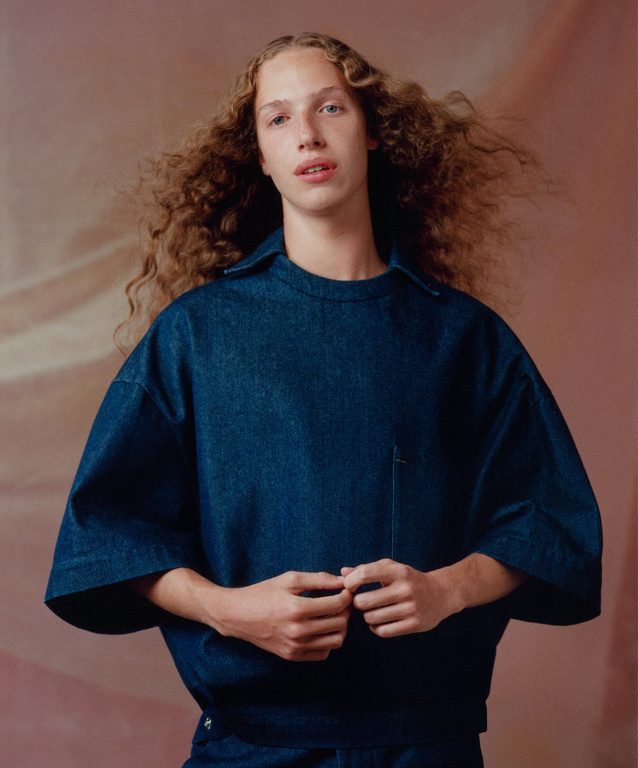 Young adult poses in denim shirt with curly hair against warm-toned backdrop in a studio portrait promoting fashion and textile textures.