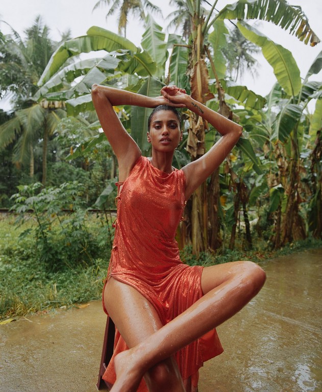 A tanned adult female poses wet in an orange dress, seated on a chair outdoors surrounded by tropical plants in the background.