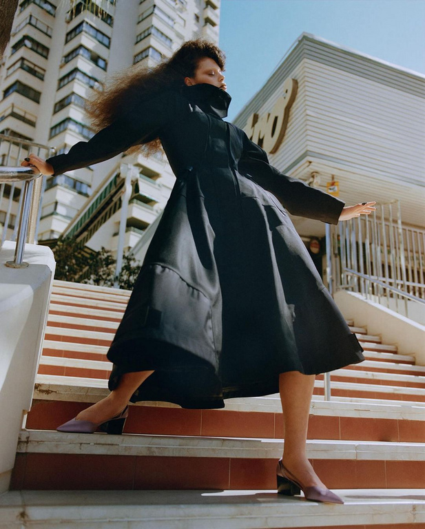 A woman in a black coat poses on outdoor steps with a modern building backdrop in a fashion editorial shot, showcasing stylish clothing.