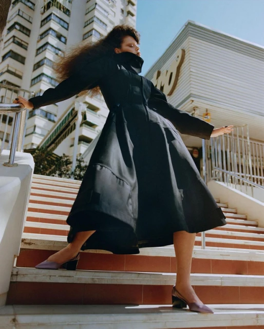 A woman in a black coat poses on outdoor steps with a modern building backdrop in a fashion editorial shot, showcasing stylish clothing.