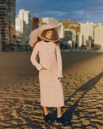 Model wearing a pink long coat and large tulle veiled hat standing on a sandy beach with city buildings in the background.
