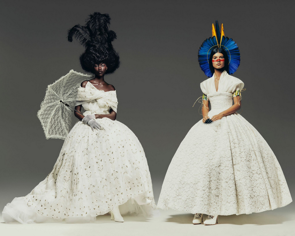Two fashion models in voluminous, white Victorian-inspired lace dresses pose in a photo studio. One holds a lace parasol, and the other wears a large feather headdress.