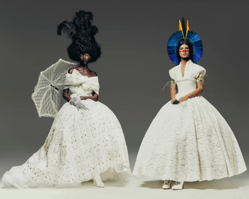 Two fashion models in voluminous, white Victorian-inspired lace dresses pose in a photo studio. One holds a lace parasol, and the other wears a large feather headdress.
