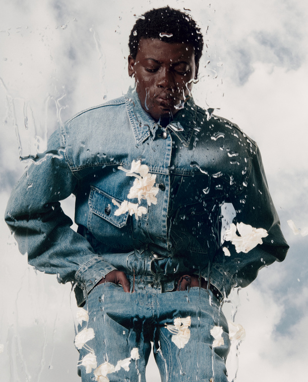 Portrait of a Black man in a denim jacket and jeans, splattered with white paint and water drops in front of a cloudy sky.