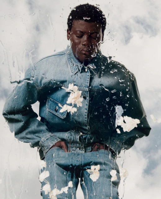 Portrait of a Black man in a denim jacket and jeans, splattered with white paint and water drops in front of a cloudy sky.