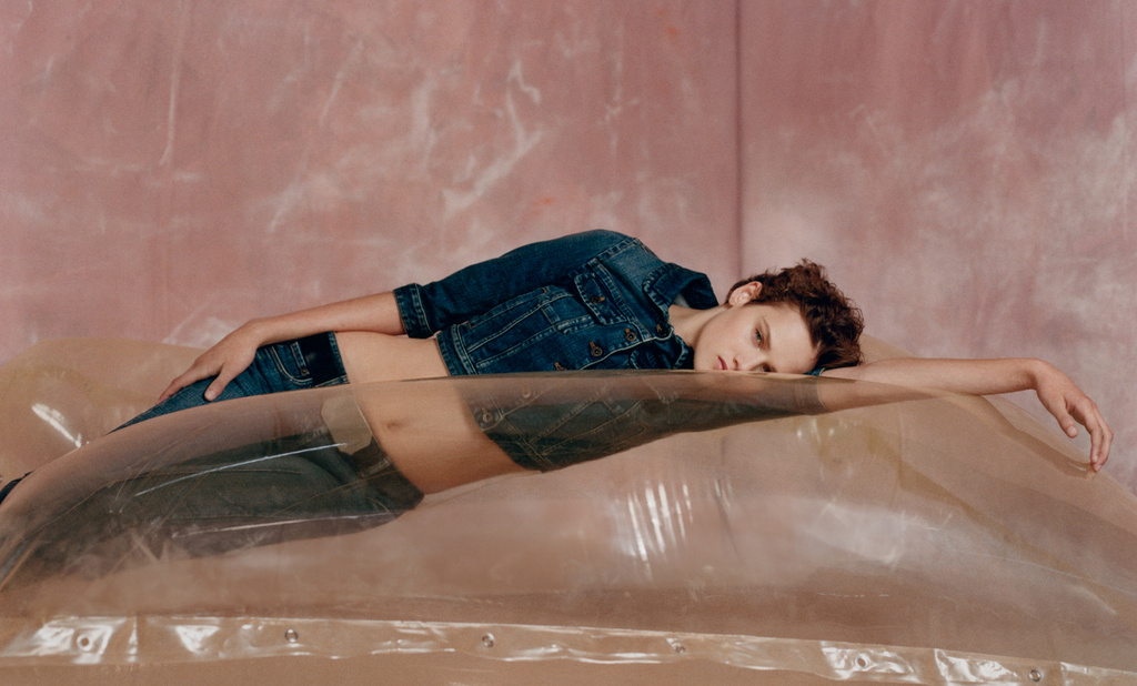 Young woman in a denim jacket and jeans lying on a transparent inflatable, posed against a pink textured wall in a fashion shot.