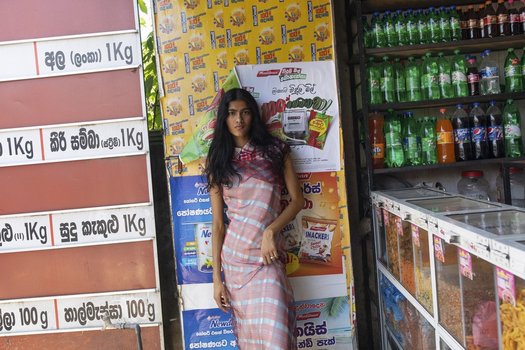 A young adult female stands in front of posters near a shop with beverage bottles and snacks visible on shelves in an eye-level medium shot.