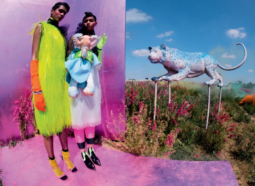 Fashion editorial featuring two young women in brightly colored, textured clothing, contrasted against a landscape with an animal sculpture.