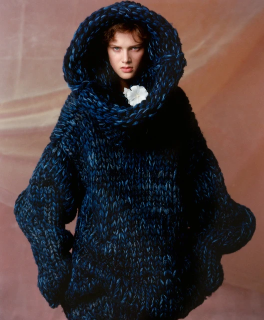 A woman poses wearing an oversized, hooded, textured, blue knit sweater against a gradient background in a fashion photography setting.