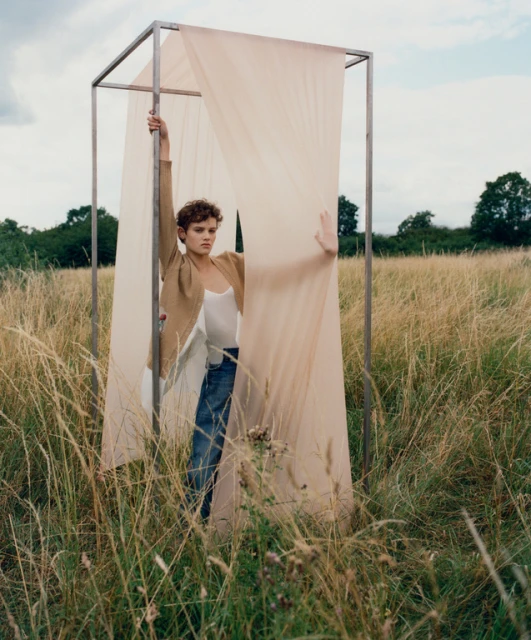 Woman in a beige blazer and blue jeans stands in a field within a metal frame draped with fabric, surrounded by tall grass and trees.