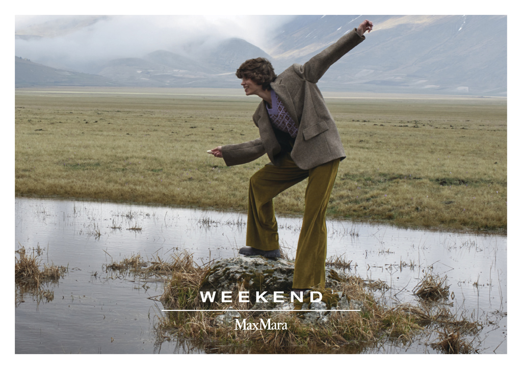 A fashion-style image showcasing a person in a brown jacket and yellow pants, standing on a rock in a flooded grassland, mountain background.