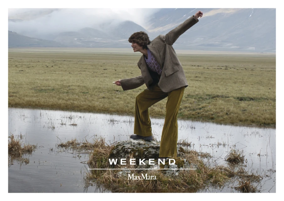 A fashion-style image showcasing a person in a brown jacket and yellow pants, standing on a rock in a flooded grassland, mountain background.