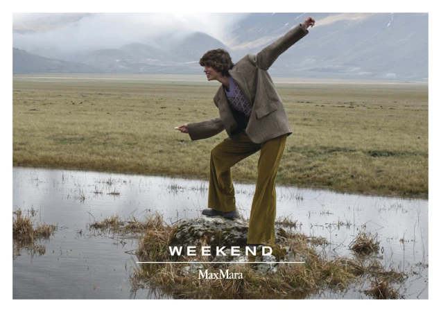 A fashion-style image showcasing a person in a brown jacket and yellow pants, standing on a rock in a flooded grassland, mountain background.