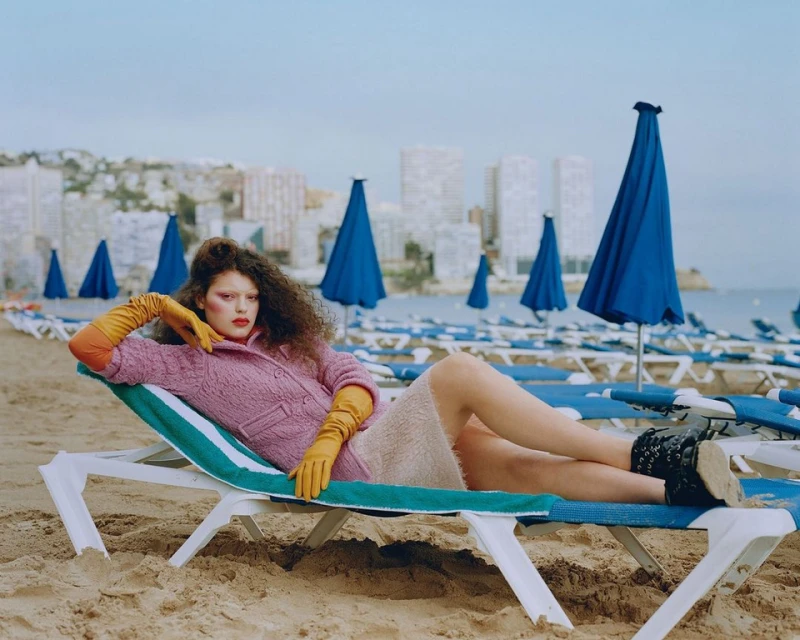 Fashion editorial shot of a woman in orange gloves reclining on a beach chair with blue umbrellas and buildings in the background.