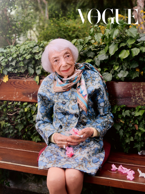 Elderly woman sits on a park bench in a floral coat with the Vogue magazine logo visible in the background amidst greenery.