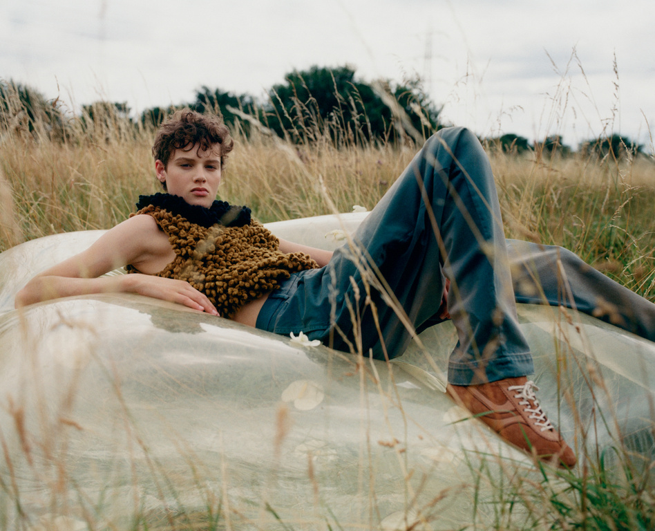 Fashionable young adult poses in a field wearing a textured top, blue jeans, and trainers, lying on a translucent surface.