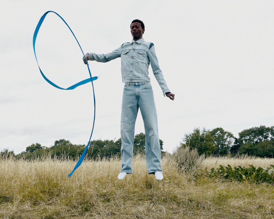 Black man wearing light wash denim jacket and jeans jumping in a grassy field while holding a bright blue ribbon.