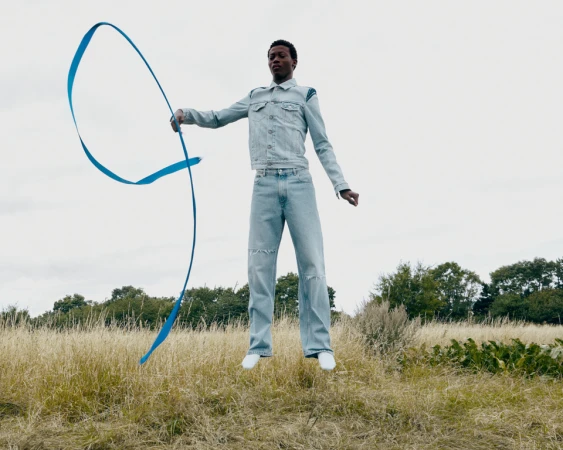 Black man wearing light wash denim jacket and jeans jumping in a grassy field while holding a bright blue ribbon.