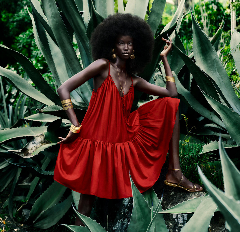 A model with dark skin and an afro wears a red dress and gold jewelry, sitting among large agave plants in a jungle-like outdoor setting.