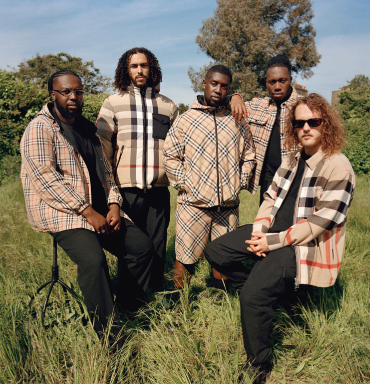 Five adult male models pose in Burberry plaid jackets and black pants outdoors in a grassy field under a blue sky with a few trees.