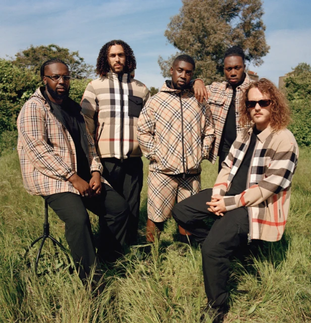 Five adult male models pose in Burberry plaid jackets and black pants outdoors in a grassy field under a blue sky with a few trees.