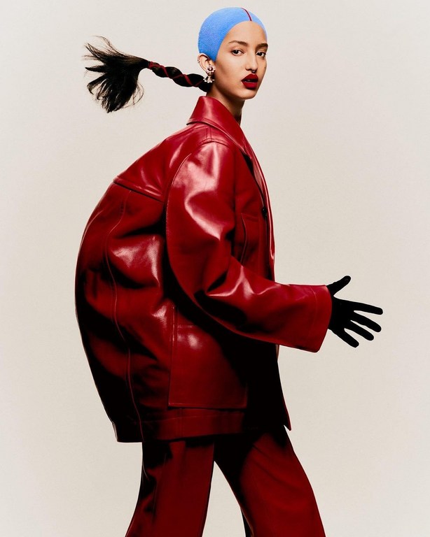 A model in a red, oversized leather suit, wearing a blue swim cap and black gloves, walks against a neutral backdrop in a fashion editorial.
